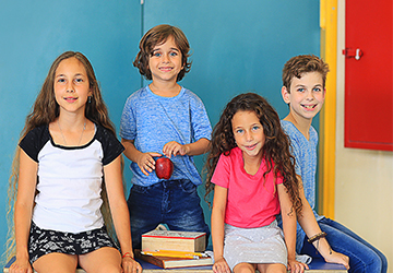 ארבעה ילדים יושבים לובשים חולצות בית ספר ילדה לובשת חולצה לבנה שרוול קצר אפור ילד חולצה כחולה ילדה חולצה ורודה ועוד ילד עם חולצה כחולה יושבים על שולחן בכיתה