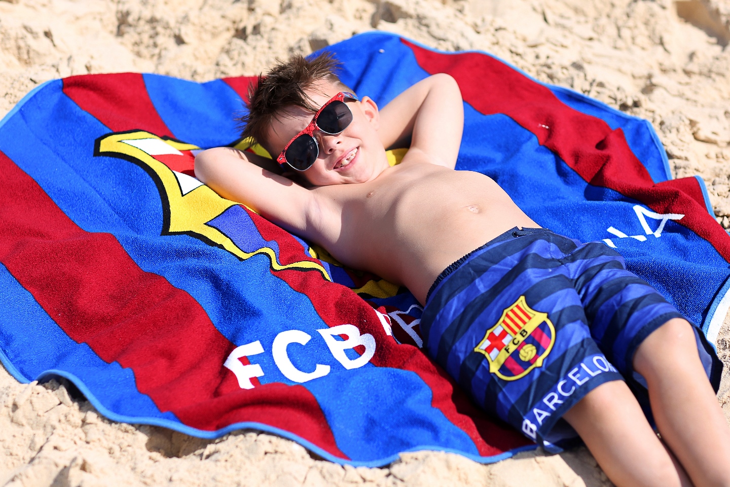 ילד שוכב בחוף ים על חול ומגבת של קבוצת כדורגל בקצלונה צבעים כחול ובורדו הילד לובש מכנס גלישה של ברצלונה בצבע כחול