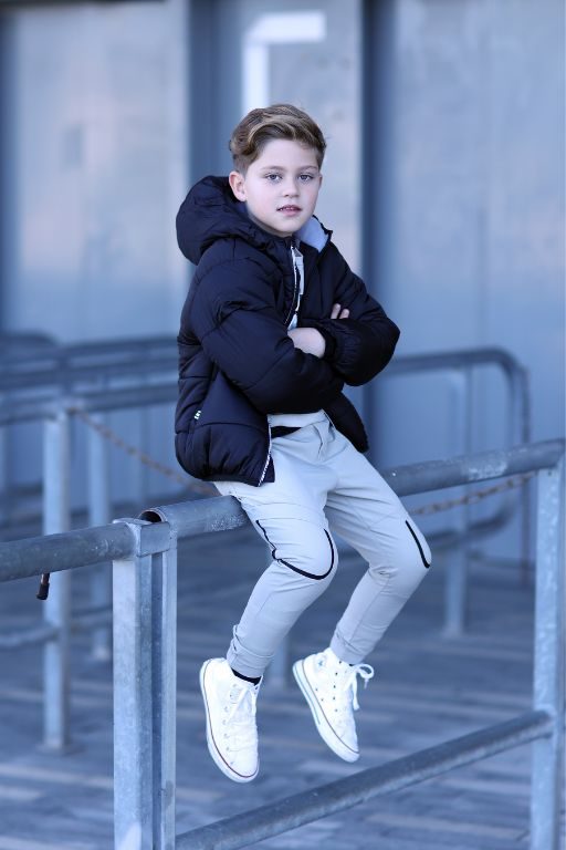ילד יושב על גדר לובש מעיל שחור ומכנסיים ארוכים אפורים